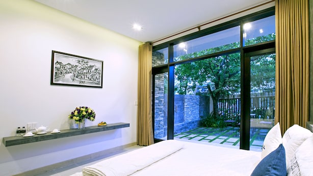 Habitación de hotel moderna con una cama grande, vistas a un jardín a través de un ventanal corredero, un cuadro en la pared y un pequeño estante con artículos de aseo.