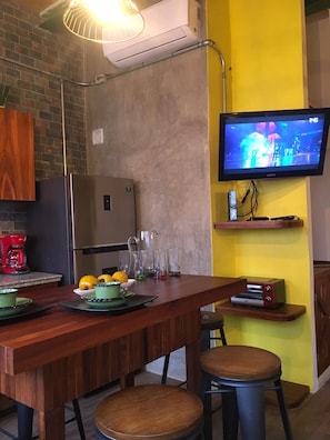 Une cuisine avec une table en bois, deux tabourets, un réfrigérateur, une machine à café et un téléviseur fixé au mur.