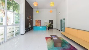 Un hall d’entrée moderne avec un comptoir de réception, des lampes suspendues aux couleurs vives, une grande fenêtre donnant sur des palmiers et un tapis coloré.
