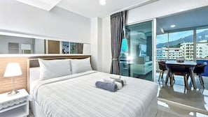 Ein Hotelzimmer mit einem Bett, einem Nachttisch, einer Lampe, einem Sessel, einem Tisch und Blick auf Gebäude durch ein Fenster.
