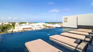 An infinity pool with lounge chairs, a clear sky, and a view of buildings and the ocean. - It Residence, 2 Bedroom Apartment with Rooftop Pool (Playa del Carmen)