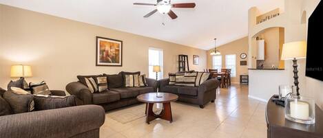 A spacious living room with two sofas, a coffee table, and a ceiling fan. A kitchen area is visible in the background.