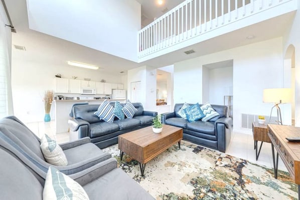 A spacious living area with a modern kitchen in the background, a balcony with a railing, and a coffee table with decorative pillows.