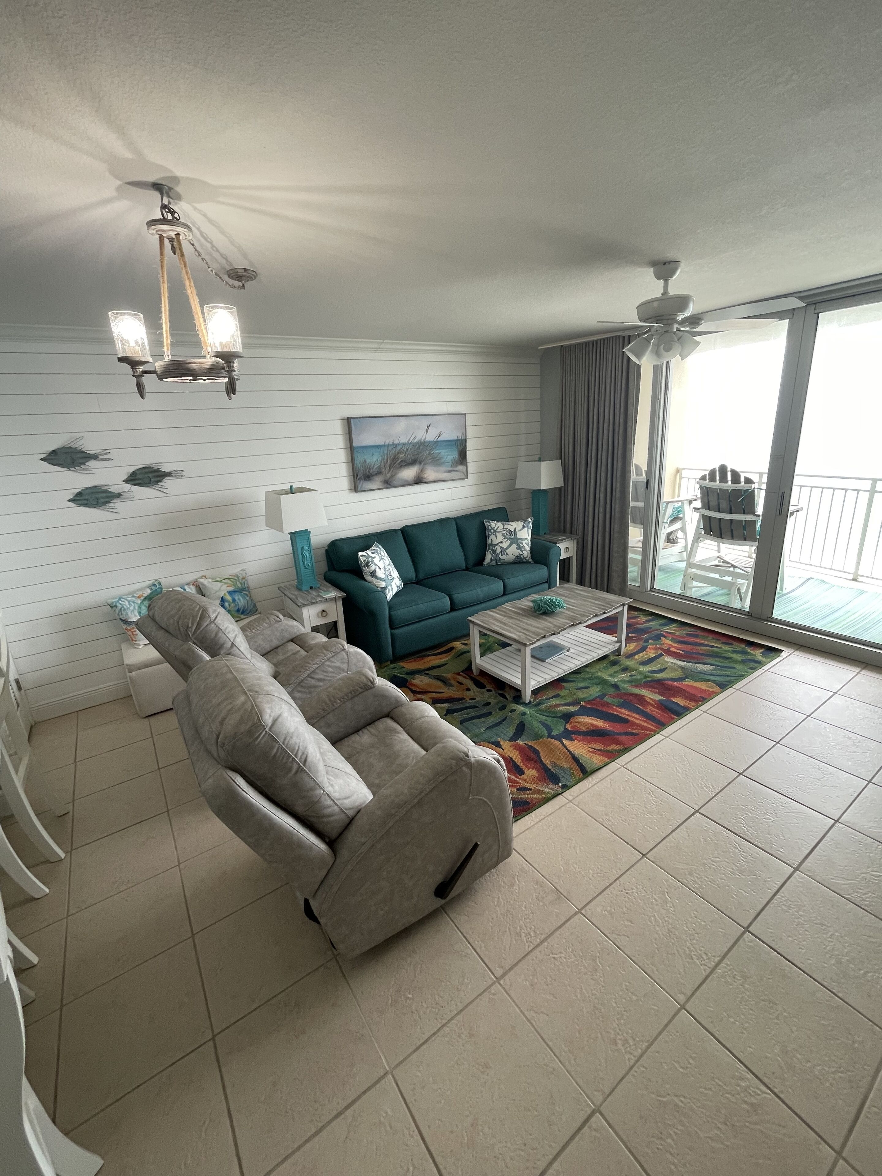 A living room with a sofa, a coffee table, a reclining chair, a ceiling fan, and a view of the outside balcony.