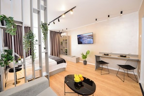 A modern, well-lit living room with a sofa, a dining table with chairs, a TV mounted on the wall, and potted plants.