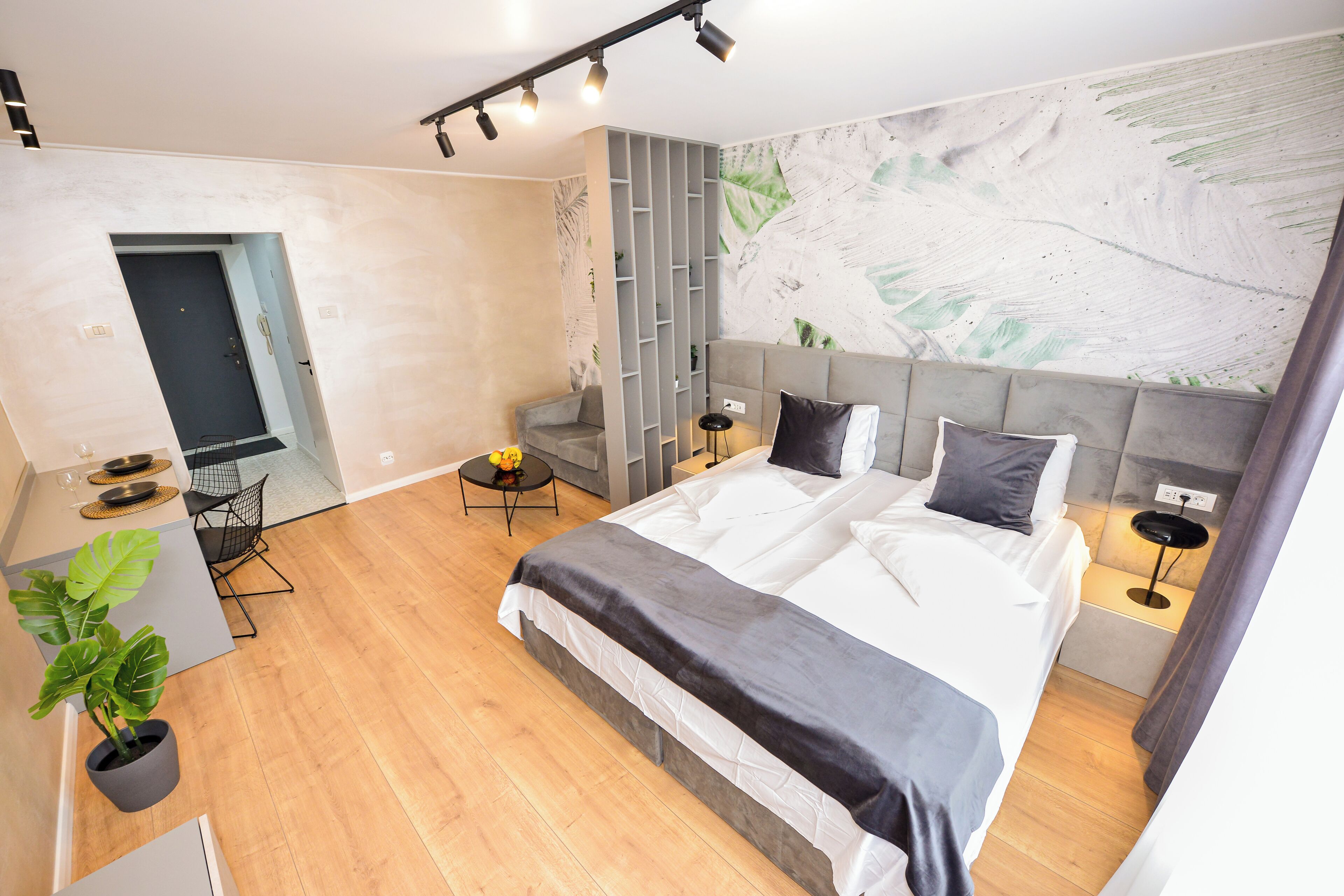 A modern hotel room with a large bed, a desk, a chair, a small table with a plant, and a wall with a leaf pattern.