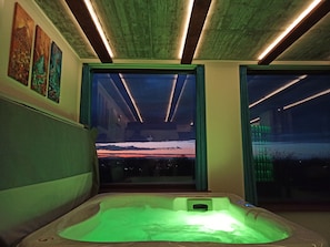 A spa room with a hot tub, wooden ceiling beams, and a view of a sunset through a large window.
