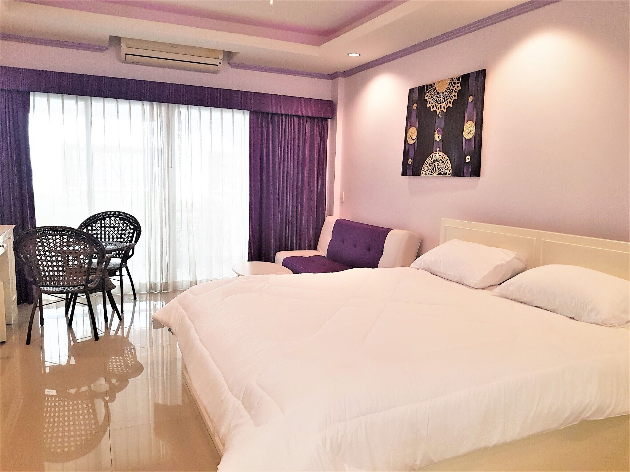 Une chambre d’hôtel avec un grand lit, deux fauteuils, une petite table, un canapé violet et un élément décoratif mural.