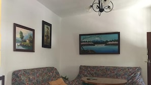 Een woonkamer met een bank en een ronde tafel, twee ingelijste schilderijen aan de muur, en een kroonluchter.