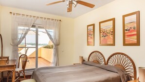 Ein Schlafzimmer mit Bett, Stuhl, Tisch und Deckenventilator. An der Wand hängen drei Gemälde.
