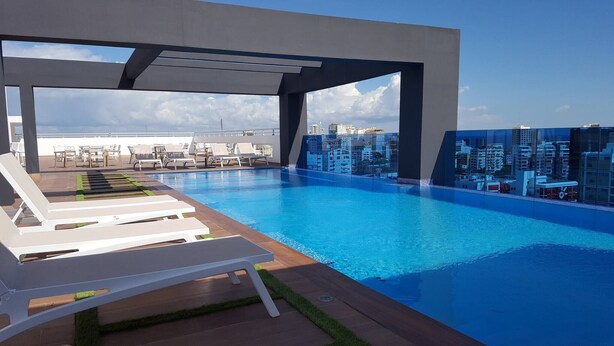 A rooftop pool with white lounge chairs, a glass barrier, and a cityscape view. - Apartment Of 1 Bedroom Plus Study With Sofa Bed (Santo Domingo)