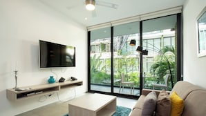 A modern living room with a flat-screen TV, a wooden coffee table, a beige sofa with cushions, and a large sliding glass door leading to a balcony with plants and a view of a building. - Lovely One Bedroom With Amazing Terrace View!! (Playa del Carmen)