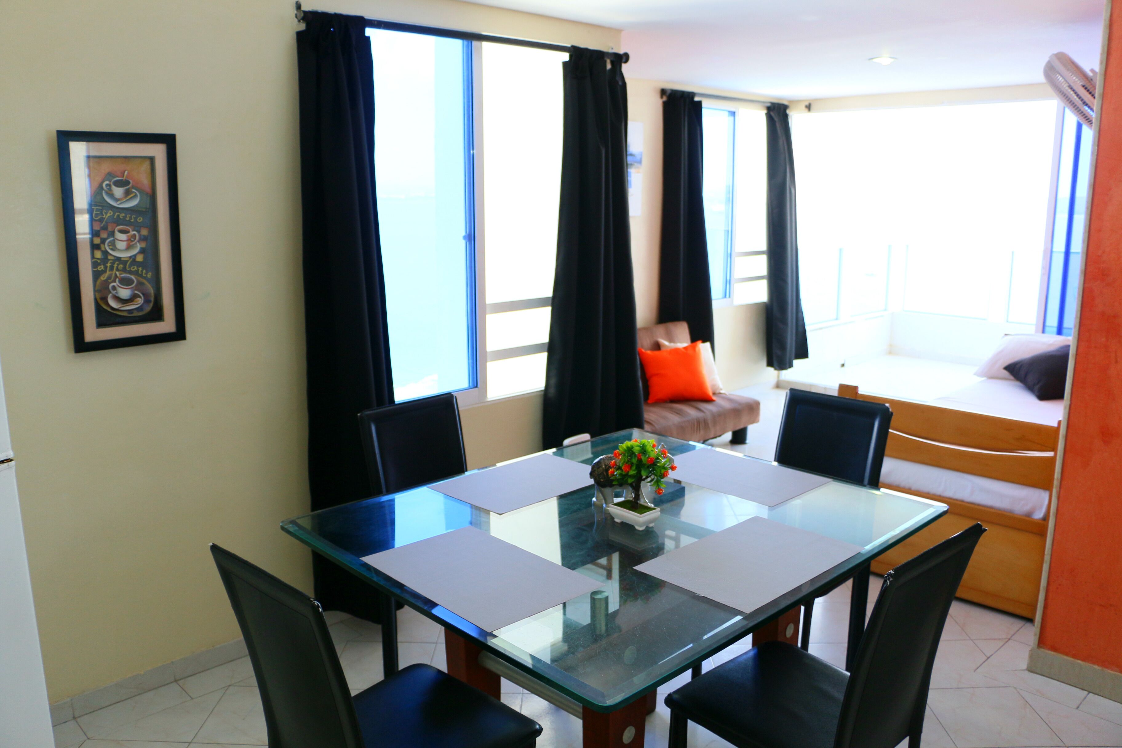 A dining area with a glass table and chairs, a small plant centerpiece, and a sofa with an orange pillow.