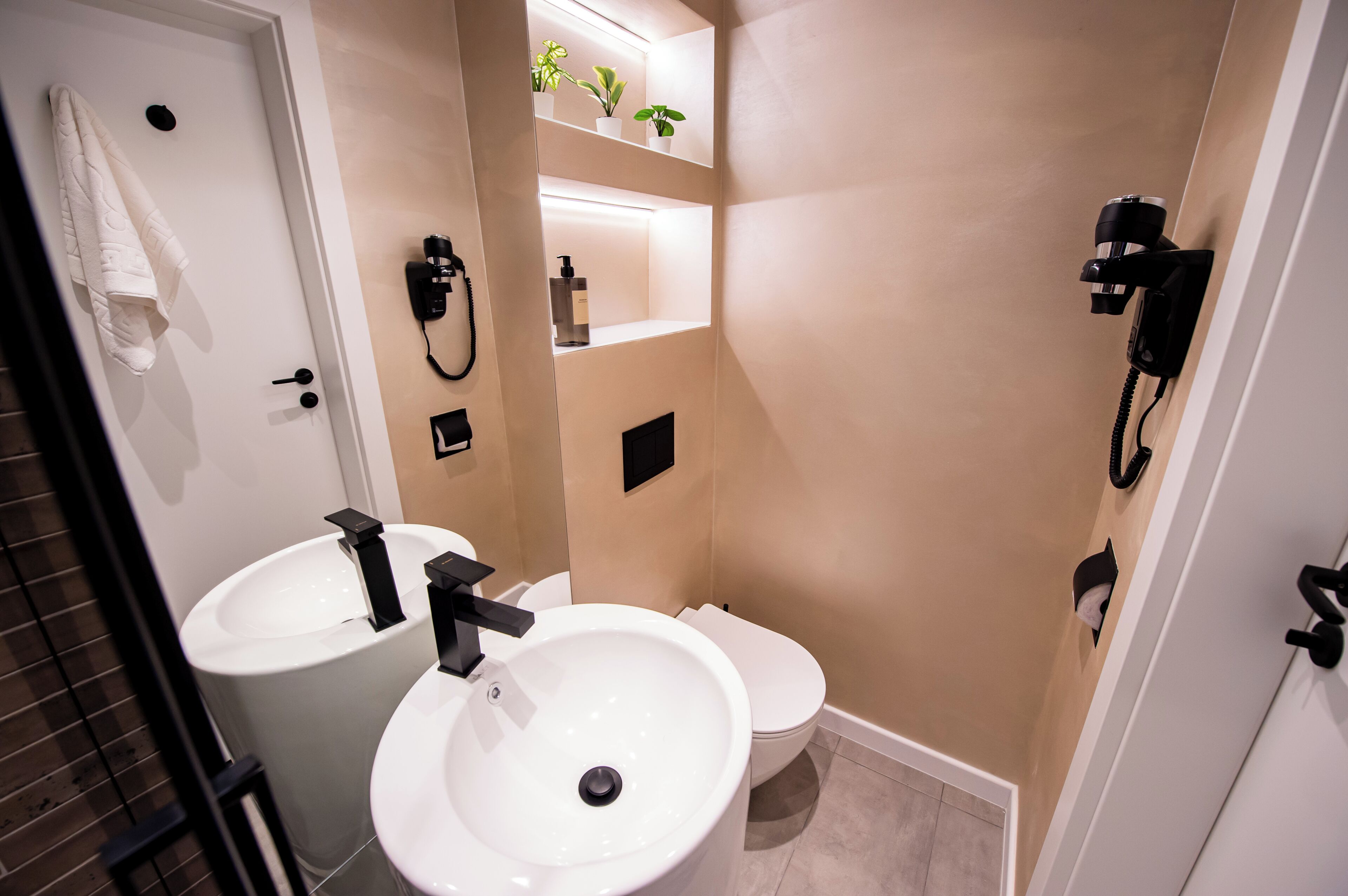 A bathroom with two sinks, a toilet, and a shower. There are plants on a shelf above the toilet.