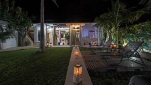 A well-lit outdoor area with a pathway leading to a building entrance, surrounded by palm trees and a wooden deck with lounge chairs. - Villa Splantzia-heated Pool-garden-2 min Chania Port (Chania)