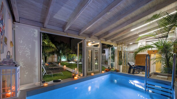 An indoor swimming pool with a glass enclosure, a wooden ceiling, and a view of a garden with outdoor seating and plants. - Villa Splantzia-heated Pool-garden-2 min Chania Port (Chania)