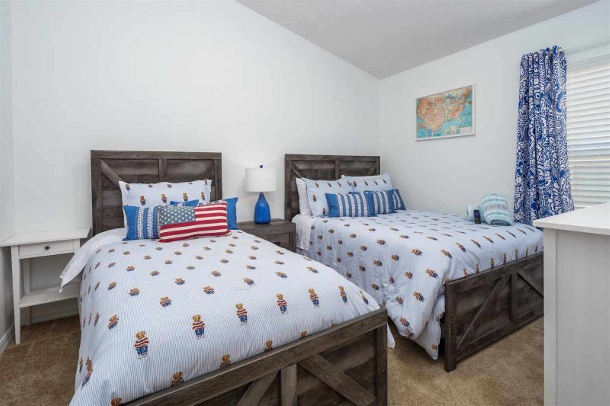 Een slaapkamer met twee bedden, elk met een houten hoofdbord, wit beddengoed met een patroon en verschillende kussens. Tussen de bedden staat een klein tafeltje met een lamp, en aan de muur hangt een kaart.