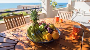 A round wooden table with a fruit bowl containing bananas, apples, grapes, and oranges, and two orange cups on a balcony overlooking the sea. - Sea Front Apartment 5 Beds With Balcony 250 Meters From the Beach (Sciacca)