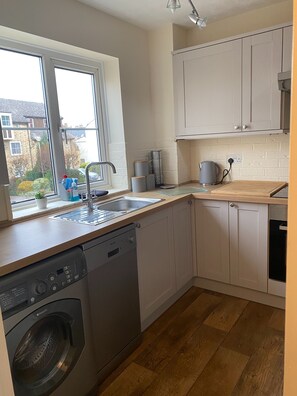 A compact kitchen with a dishwasher, a washing machine, a window with a view of houses, and a kettle on the counter. - Waterfront House 3 BR Parking St Neots Town Centre (St Neots)