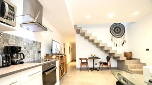 Cozinha moderna com micro-ondas, máquina de café e forno embutidos. Possui um espaço de jantar com mesa de vidro e cadeiras. Uma escada decorada com um sonho indígena dá acesso a um andar superior.