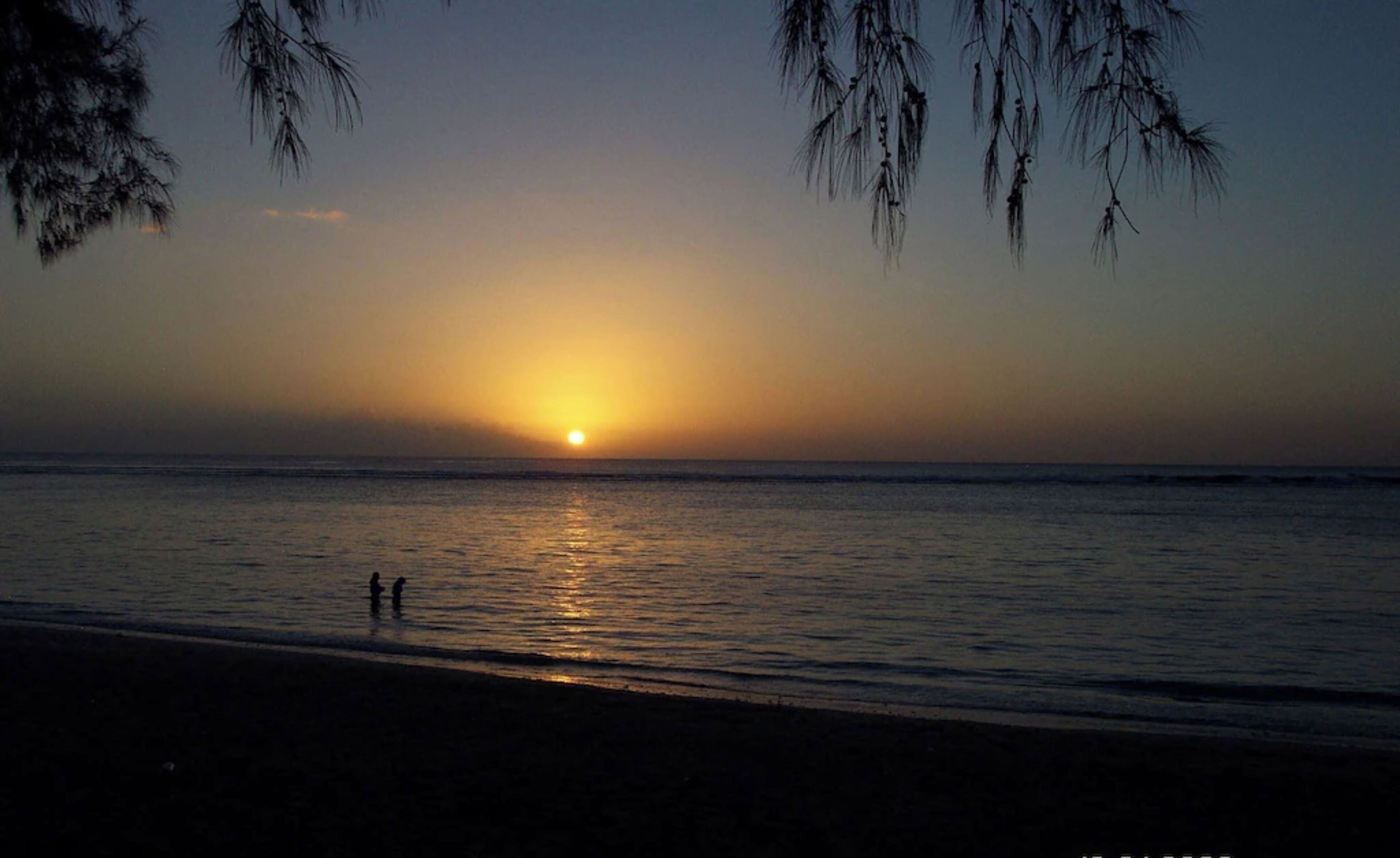 Un coucher de soleil sur une mer calme, avec les silhouettes de personnes sur la plage.