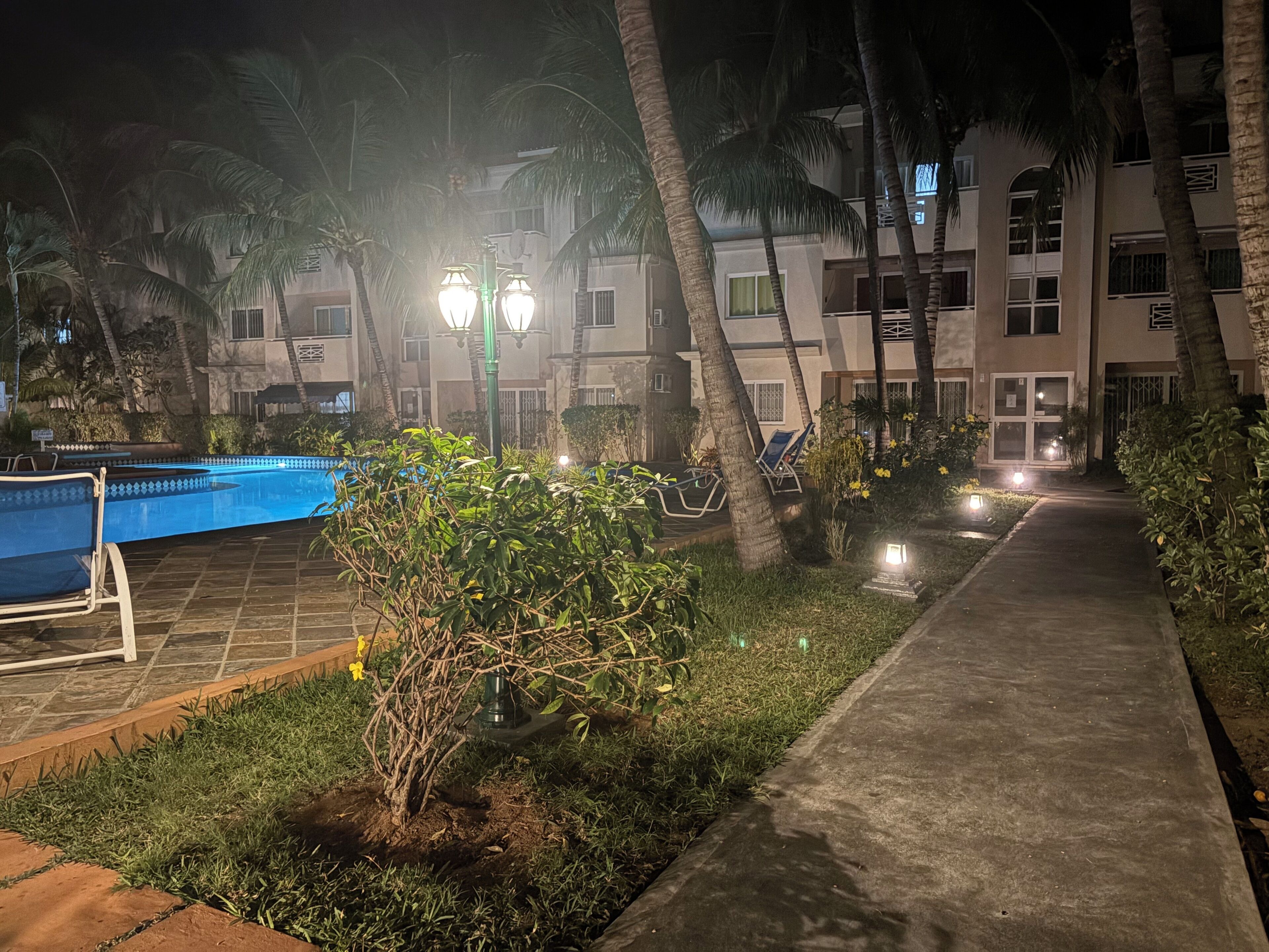 Vista nocturna de un área de piscina de hotel, con piscina, sillones de descanso, palmeras y un camino que lleva al edificio.