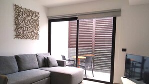 A modern living room with a grey sectional sofa, a flat-screen TV, and a sliding glass door leading to an outdoor area with a table and chairs. - Private Roof w Plunge Pool, Brand New 2 Br Penthouse for 6 Sleeps! (Tulum)