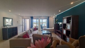 A modern living room with a sofa, armchairs, a coffee table, and a TV stand. There is a balcony with a view of the outdoors. - 88 - Livingtarifa La Marea (Tarifa)