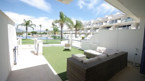 A balcony with a wicker sofa, a small table, and a chair. There is a pool in the background with palm trees and a clear sky. - 88 - Livingtarifa La Marea (Tarifa)