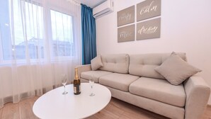 Een moderne woonkamer met een beige bank, een ronde witte salontafel en een fles champagne. Gordijnen, een airconditioning en motiverende kunst aan de muur.