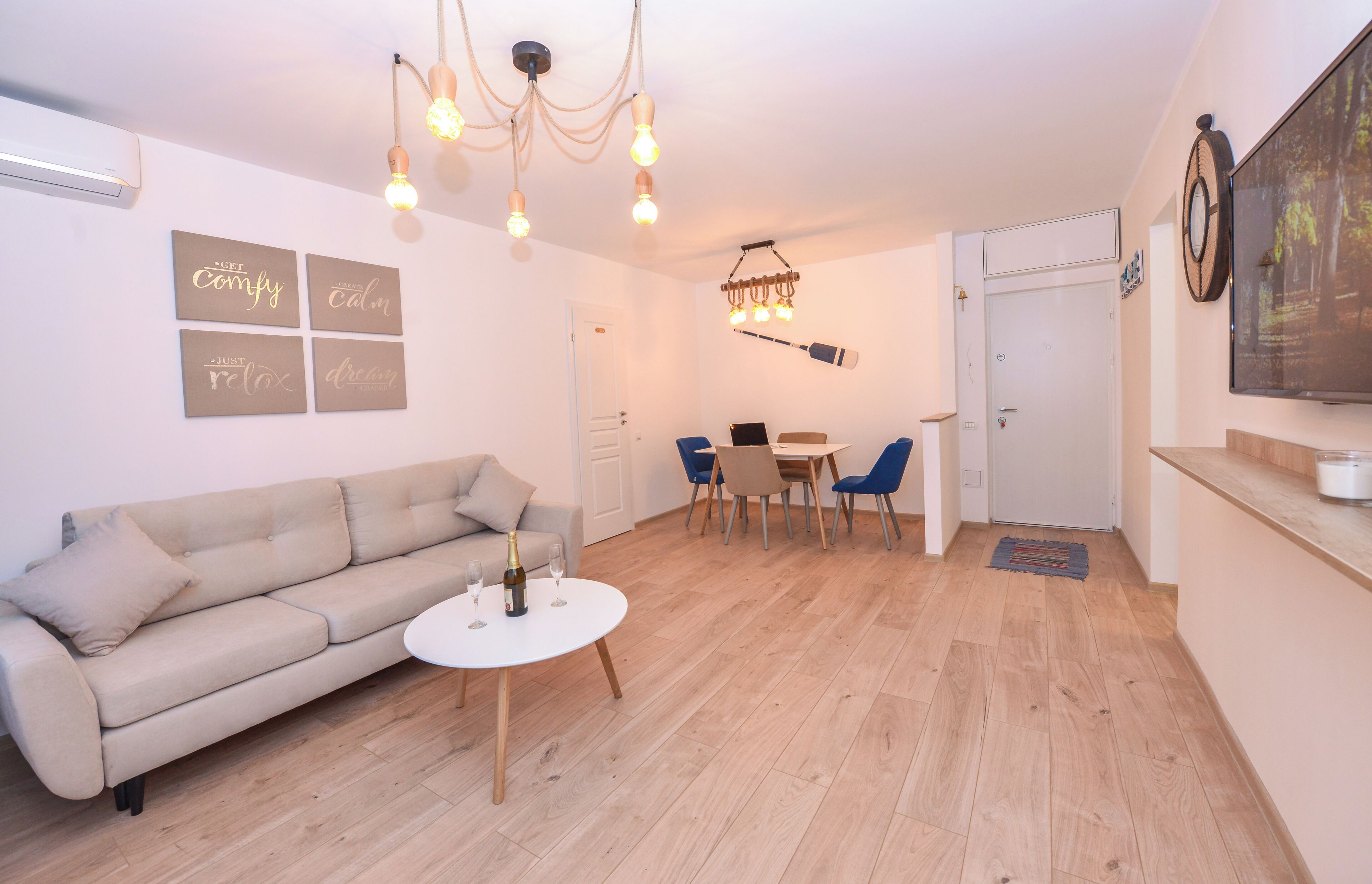 A modern living room with a sofa, a dining table, and chairs. There is a chandelier, a wall-mounted TV, and a door.