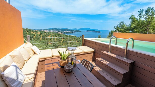 A balcony with a wooden table, a cushioned seating area, and a view of a body of water and greenery. - Villa Kallisto With Private Pool And Stunning Sea Views (Skiathos)