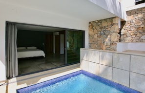 A modern hotel room with a glass door leading to a balcony, a stone wall, and a small pool area.