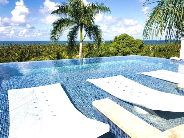 A clear blue infinity pool with white lounge chairs, surrounded by lush greenery and a view of the ocean.