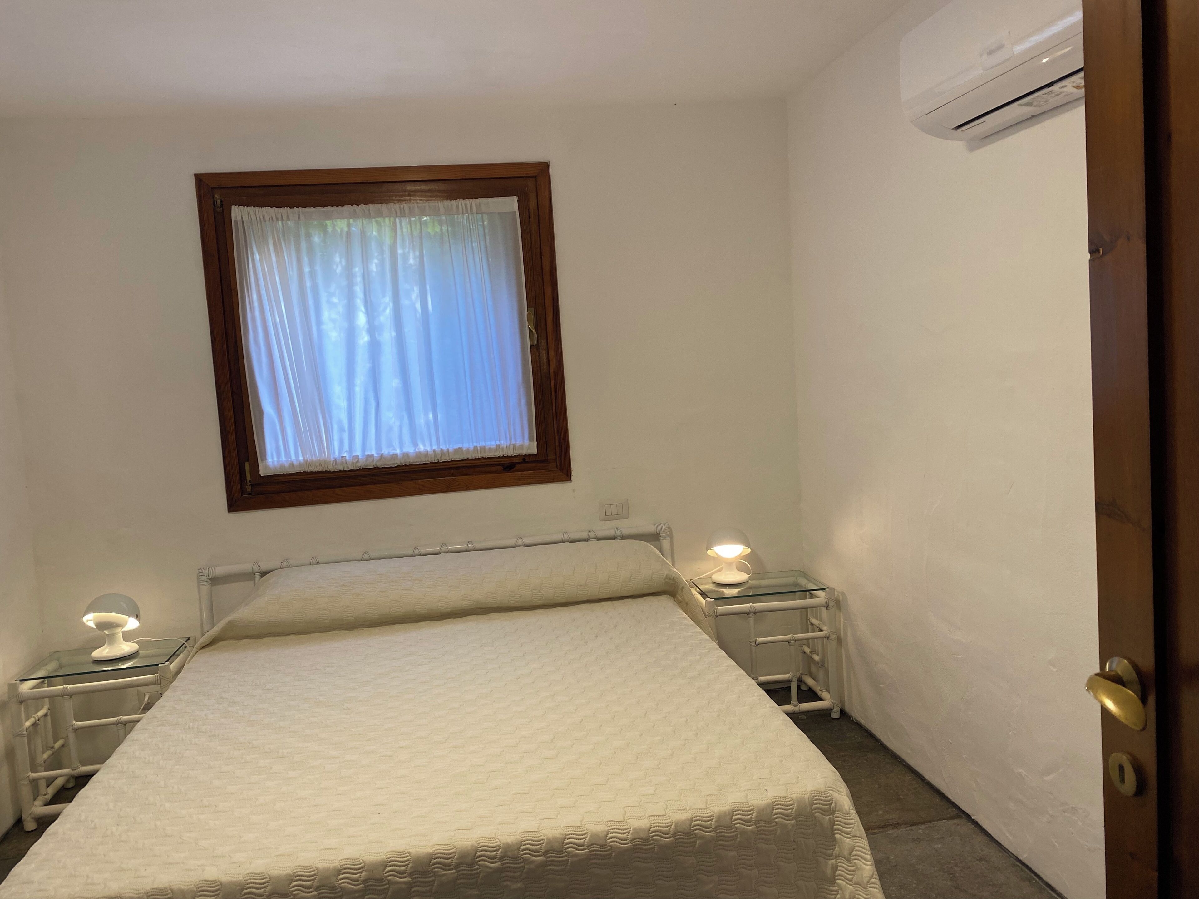 A small, neatly arranged bedroom with a single bed, a window with curtains, a bedside lamp, and an air conditioning unit.
