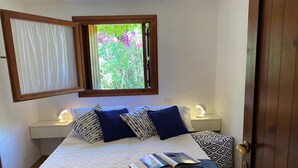 A neatly made bed with a patterned pillow, a bedside lamp, and a window with a view of greenery. - Domus Olivarum -costa Smeralda Villetta 7 Persone, 4 PK, Wifi, Grande Giardino (Arzachena)