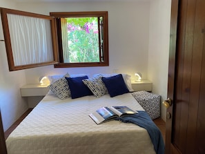 Un lit bien fait, agrémenté d’un coussin à motifs, d’une lampe de chevet et d’une fenêtre donnant sur un paysage verdoyant.