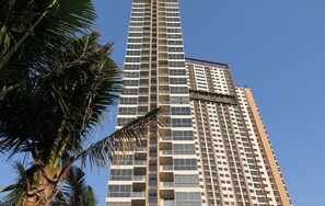 A tall, modern high-rise building with multiple floors and balconies, surrounded by palm trees.
