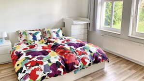 Een slaapkamer met een kleurrijk bloemenbedovertrek, een witte nachtkastje met een lamp en een raam met gordijnen.
