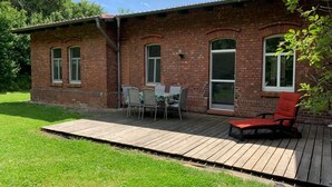 Een bakstenen huis met een houten terras, een eettafel met stoelen, en een rode stoel.