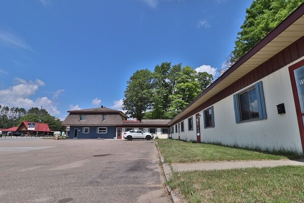 A motel with a single-story structure, a parking lot, and a red-roofed building in the background. - BestLiving Motel (Severn)