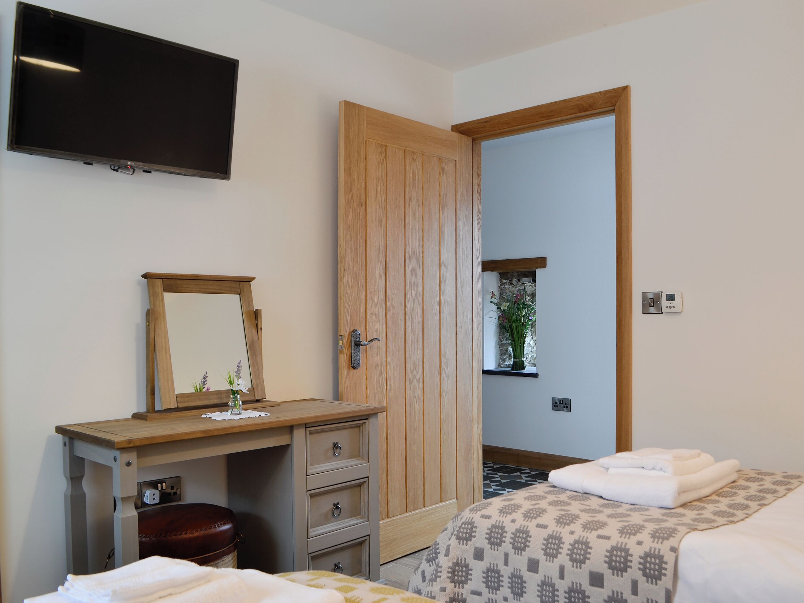Ein Hotelzimmer mit Holzschreibtisch, Fernseher, Kleiderschrank, einem Bett mit Handtüchern und einem kleinen Fenster mit Pflanze.