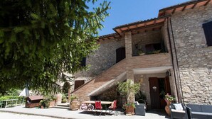 Un edificio de piedra con una escalera de madera que conduce a un balcón. Hay mobiliario exterior con sillas rojas y una mesa.