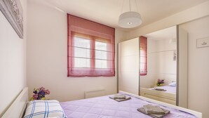 A bedroom with a bed, a window with curtains, a bedside table with a vase of flowers, and a mirror on the wall. - TIVAT apartment (NO-9) (Donja Lastva)