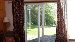 Ein Zimmer mit einer Schiebeglastür, die zu einem Garten führt. Ein Korbstuhl mit Blumenkissen und eine Lampe auf einem Holztisch sind zu sehen.