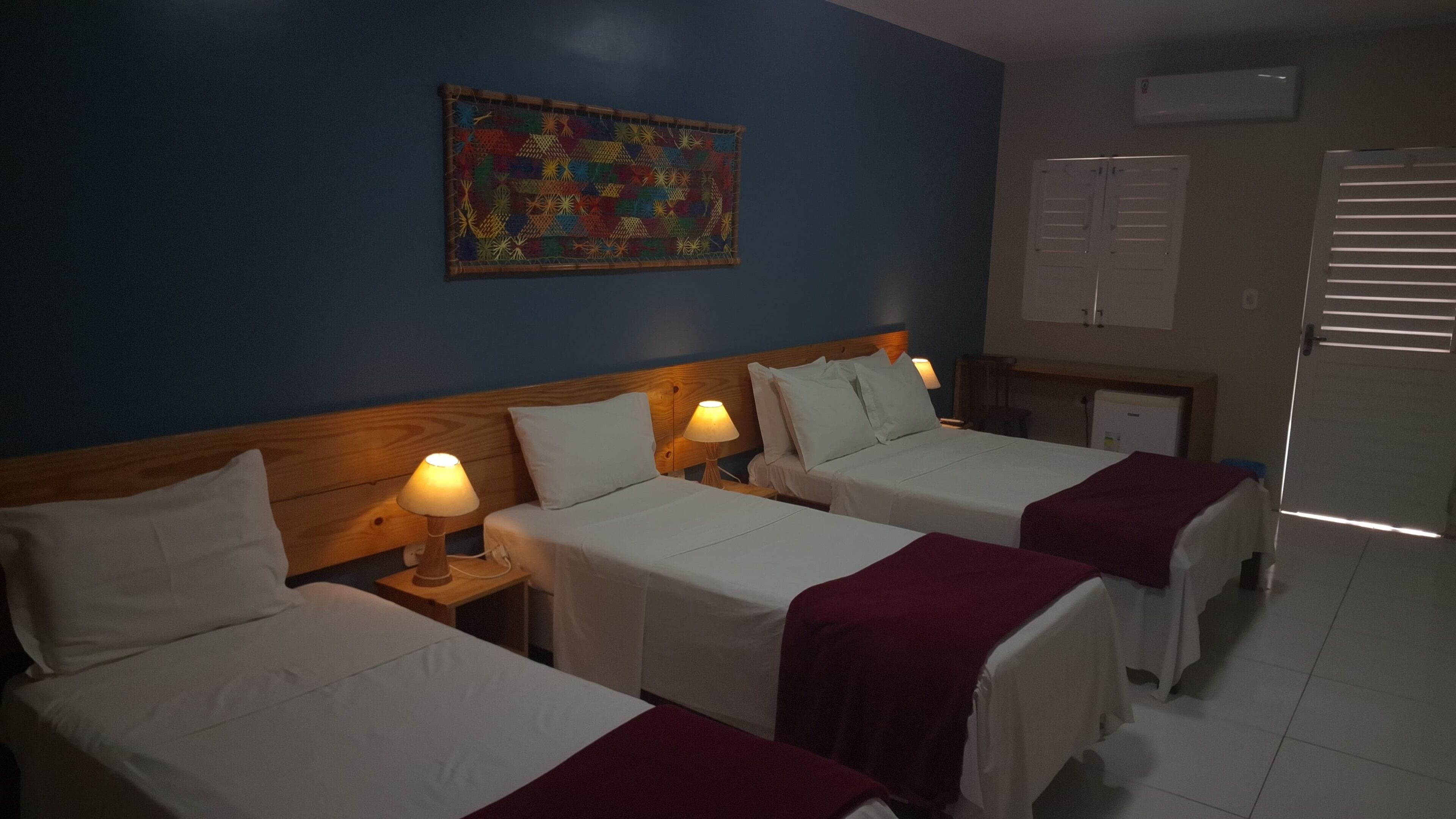 Une chambre d’hôtel avec deux lits, une tête de lit en bois, une décoration murale colorée et une fenêtre avec des stores.