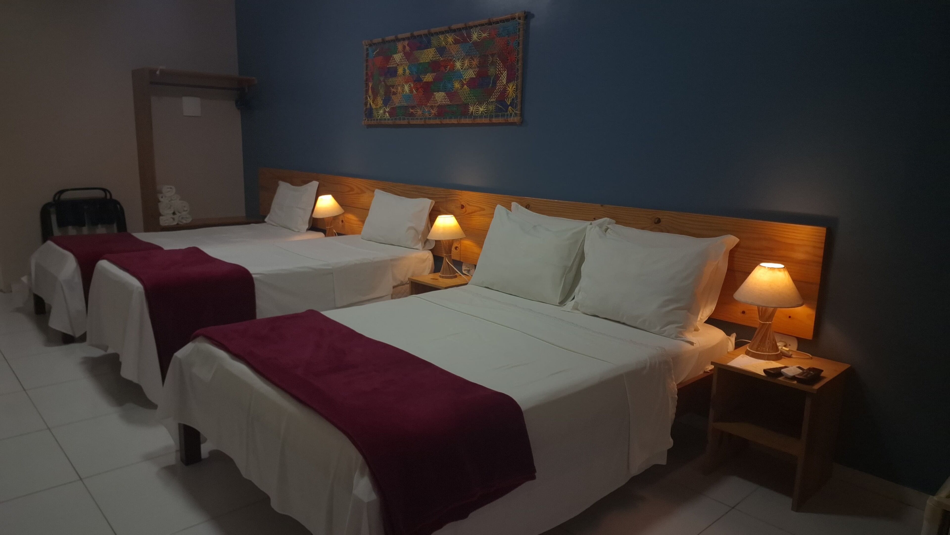 A hotel room with two beds, each with a white bedspread and a red blanket. There are bedside lamps on each nightstand. A colorful artwork is on the wall above the beds.