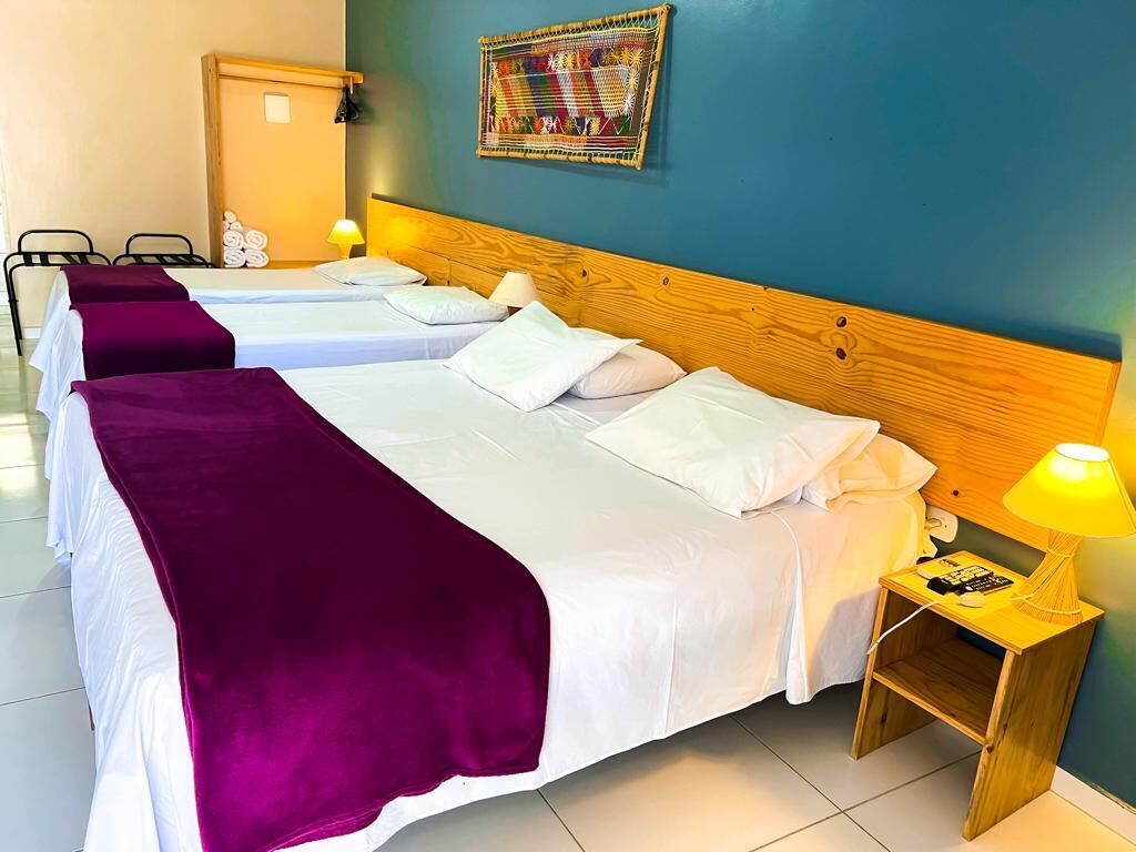 Une chambre d’hôtel avec deux lits, chacun recouvert de draps blancs et d’une couverture violette. On y trouve une tête de lit en bois, une petite table de chevet en bois avec une lampe, et une tapisserie murale colorée en fibres tissées.