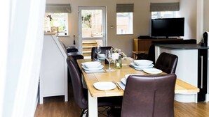 A dining area with a wooden table set for four, surrounded by leather chairs. A television is mounted on the wall to the right. - Luxury chalet in wonderful Pembrokeshire National Park, beach & coast path (Nolton Haven)