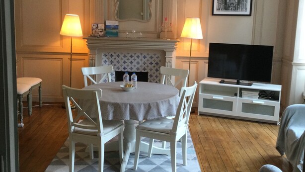 A dining area with a round table, white chairs, and a patterned rug. There is a television on a white cabinet and two lamps on either side of the table. - EGLANTINE  (Dinan)